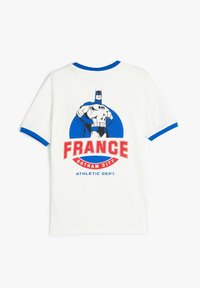 Camiseta blanca con un gran gráfico de un superhéroe con acentos azules, que dice "FRANCIA GOTHAM CITY DEPARTAMENTO ATLÉTICO" en texto rojo y azul.