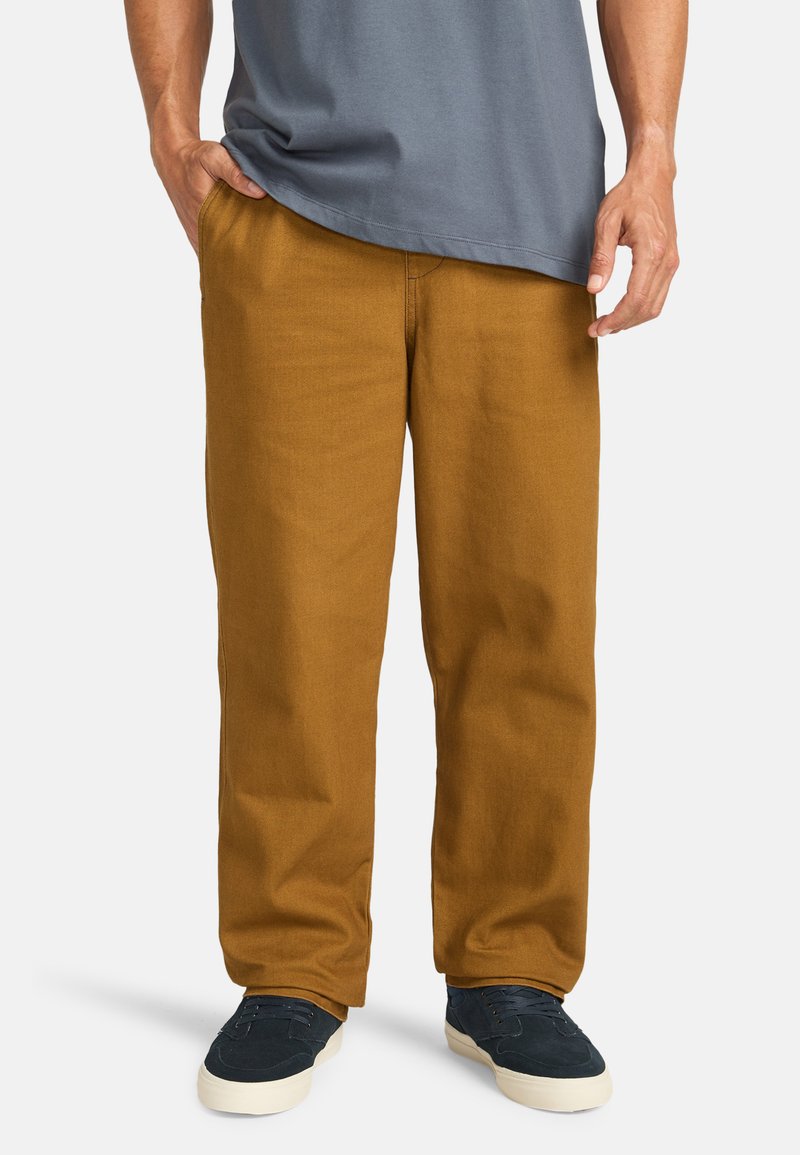 Pantaloni di cotone kaki con vestibilità rilassata, gambe dritte e una texture minimale. Abbinati a sneakers blu/neri e una maglietta grigia.
