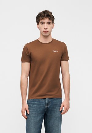 Ung mann med krøllete hår som har på seg en brun t-skjorte med korte ermer og blå jeans, står foran en ensfarget lys bakgrunn.
