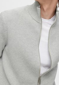 Ljusgrå zip-up-tröja med ribbade hög hals, texturerad tyg och silverdragkedja, lagrad över en vit T-shirt.