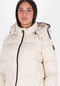 Jasnobeżowa kurtka puchowa z wysokim kołnierzem, zamek błyskawiczny z przodu oraz odczepianym kapturem. Charakteryzuje się pikowaną fakturą i czarnymi akcentami metalowymi.