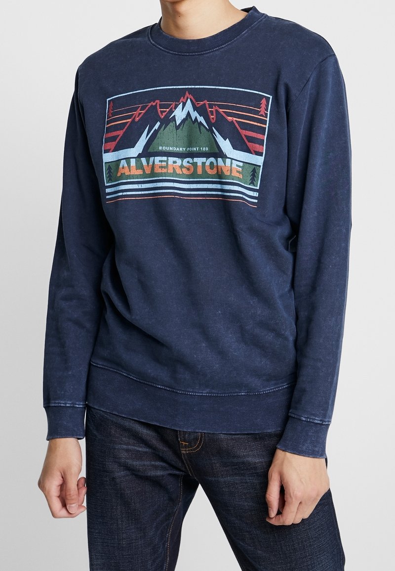 Personne portant un sweatshirt bleu marine avec un graphique de montagne et le texte "ALVERSTONE" à l'avant, associé à un jean bleu foncé.