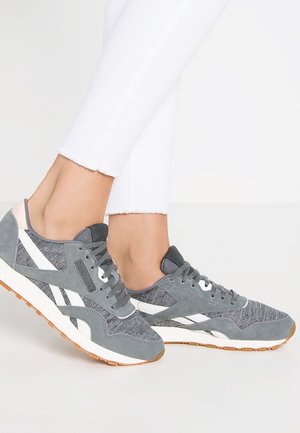 Osoba nosící šedo-bílé tenisky s gumovou podrážkou, v kombinaci s bílými zkrácenými kalhotami na jednoduchém světlém pozadí.