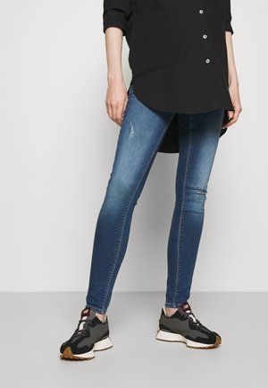 Person wearing blue skinny jeans with light distressing and black sneakers, standing against a plain light background.