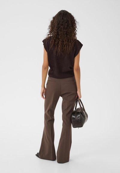 Femme aux longs cheveux bouclés se tient de dos, portant un haut sombre sans manches, un pantalon évasé marron à carreaux, et tenant un sac à main noir.