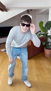 Hall halli, sinised teksad, valged tossud. Tumedad päikeseprillid hoitakse ühe käega. Siseruum, kus on taim ja pruun diivan. Puitpõrand.