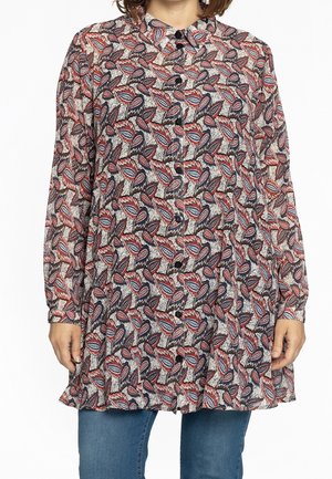 Tunique à manches longues à boutons avec un motif de feuilles rouges, bleues et blanches portée par-dessus un jean bleu, vue du cou à mi-cuisse.