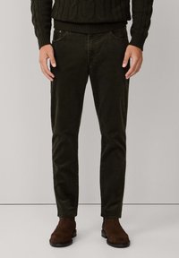 Pantalons en velours côtelé vert foncé avec cinq poches, coupe slim et texture visible. Des bottes en daim marron complètent la tenue.