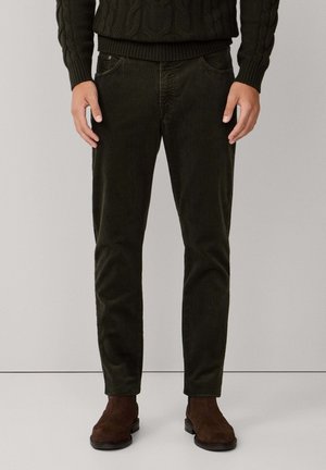Dunkelgrüne Cordhosen mit fünf Taschen, schlanker Schnitt und sichtbarer Textur. Braune Wildlederstiefel ergänzen das Outfit.