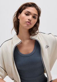 Helebeež zipiga jakk musta äärisega, kantud tumesinise soonikkoega tanki peal. Jakil on vasakul pool logo.