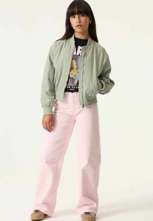 Veste aviateur vert clair avec fermeture éclair devant, pantalon rose à jambes larges, et t-shirt noir graphique avec motif floral. Baskets marron.