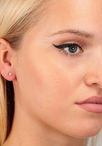 Nahaufnahme einer hellhäutigen Person mit blonden Haaren, einem kantigen Gesicht, dezenter Make-up und einem kleinen diamantbesetzten Ohrstecker im linken Ohr.