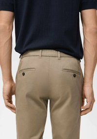 Pantaloni beige con una texture liscia e una vestibilità sartoriale. Presentano due tasche posteriori con chiusure a bottone. Indossati con una camicia blu navy.