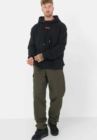 Zwarte hoodie met verstelbare koordjes en oranje logo, gecombineerd met olijfgroene cargo-broeken en zwarte sneakers. Casual, losse pasvorm.
