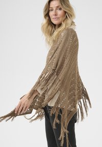 Châle en crochet marron, avec un tissage ouvert et de longues franges, drapé librement, complétant un haut de couleur claire et un pantalon sombre.