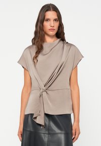 Top beige a maniche corte con colletto alto, caratterizzato da un dettaglio a nodo torcigliato in vita, realizzato in un tessuto morbido e fluido.