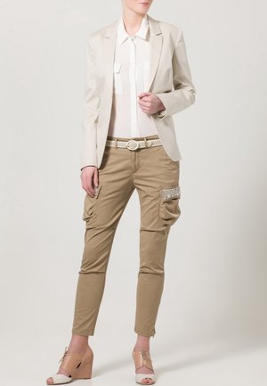 Blazer beige sur une blouse blanche, pantalon cargo beige avec poches latérales, et talons compensés marron clair avec un détail en nœud. Accents texturés sur le pantalon.