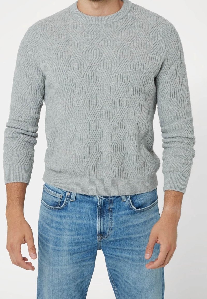 Homme portant un pull en maille texturé gris clair et un jean en denim bleu sur un fond blanc uni.
