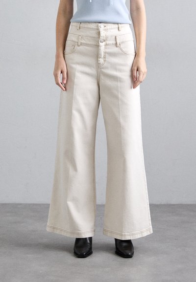 Pantalons en denim beige à jambe large avec une taille haute, cinq poches, fermeture à double bouton à l'avant, et couture contrastante, associés à des bottes à talons noires.
