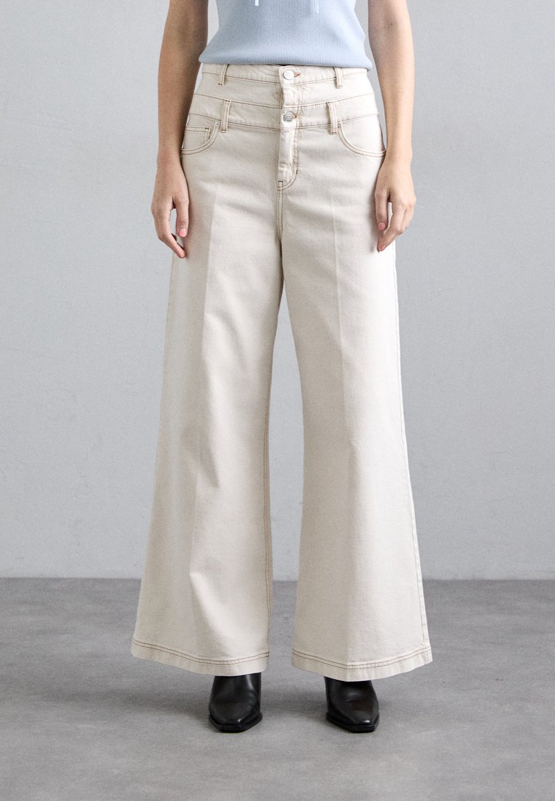 Beżowe dżinsowe spodnie o szerokich nogawkach z wysokim stanem, pięcioma kieszeniami, podwójnym zapięciem na guziki z przodu oraz kontrastowym ściegem, połączone z czarnymi botkami na obcasie.