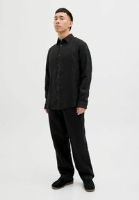 Camisa negra de botones con cuello y mangas largas, combinada con pantalones negros de corte suelto y zapatos oscuros, contra un fondo liso.