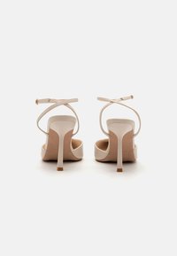 Sandales à talons beiges avec des fines lanières se croisant à l'arrière, présentées de dos sur un fond blanc.