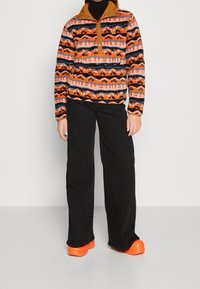 Miękki sweter z wzorem w kolorach pomarańczowym, różowym i czarnym, kołnierz w kolorze brązowym, zestawiony z czarnymi spodniami o szerokich nogawkach i pomarańczowymi butami.