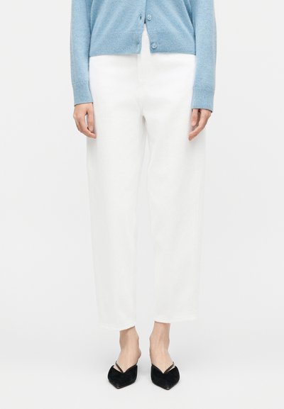 Άτομο που φοράει λευκό παντελόνι με ίσια γραμμή, ανοιχτό μπλε πουλόβερ με κουμπιά και μαύρα παπούτσια με μυτερή μύτη και χαμηλό τακούνι, στέκεται μπροστά σε λευκό φόντο.