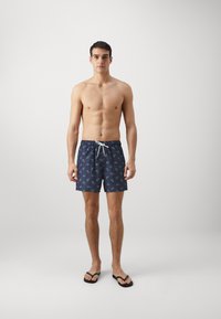 Marine svømmedrakter med et grønt bladmønster, elastisk midjebånd og hvit snor. Brukt med svarte flip-flops.