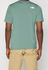 Mężczyzna w zielonej koszulce z krótkim rękawem The North Face i beżowych spodniach, pokazany od tyłu na jednolitym tle.