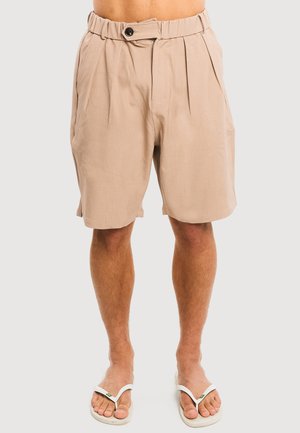 Short beige plissé taille haute avec bouton et fermeture éclair, porté par une personne aux jambes nues en tongs blanches, debout sur un fond uni.