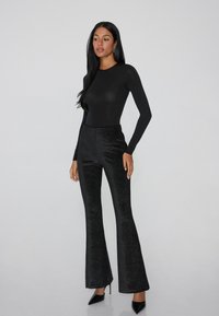 Zwarte body met lange mouwen gecombineerd met zwarte wijde broeken met een gestructureerd patroon. Model draagt puntige zwarte hakken.