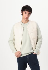 Chaleco acolchado de color beige claro con un bolsillo frontal y textura suave, superpuesto sobre un suéter verde pálido, combinado con jeans oscuros.