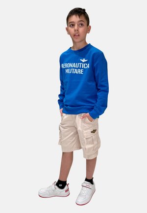 Felpa blu con stampa "AERONAUTICA MILITARE", pantaloni cargo beige con tasche laterali e sneakers bianche con dettagli rossi.