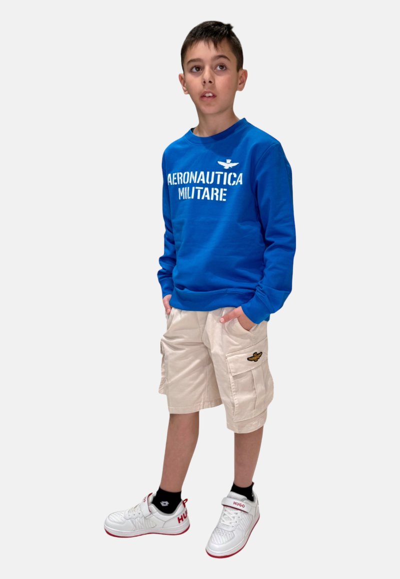 Felpa blu con stampa "AERONAUTICA MILITARE", pantaloni cargo beige con tasche laterali e sneakers bianche con dettagli rossi.
