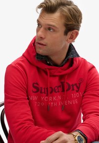 Czerwona bluza z kapturem z logo na przedzie, sznurkami oraz kołnierzykiem szarej koszuli w kratę. Widoczny zegarek na nadgarstku z niebieskim paskiem.