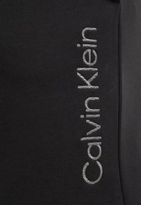 Tejido negro con el nombre bordado "Calvin Klein" en hilo plateado, situado verticalmente a lo largo de un lado. Textura suave.