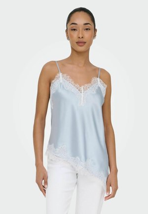 Kobieta ubrana w jasno niebieską satynową koszulkę na ramiączkach z białą koronkową lamówką i białe spodnie, stojąca na jednolitym jasnym tle.