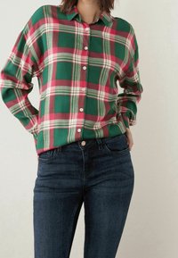 Chemise à carreaux verts et rouges à boutons avec col ; comporte des boutons blancs et des manches retroussées. Associée à un jean slim bleu marine.