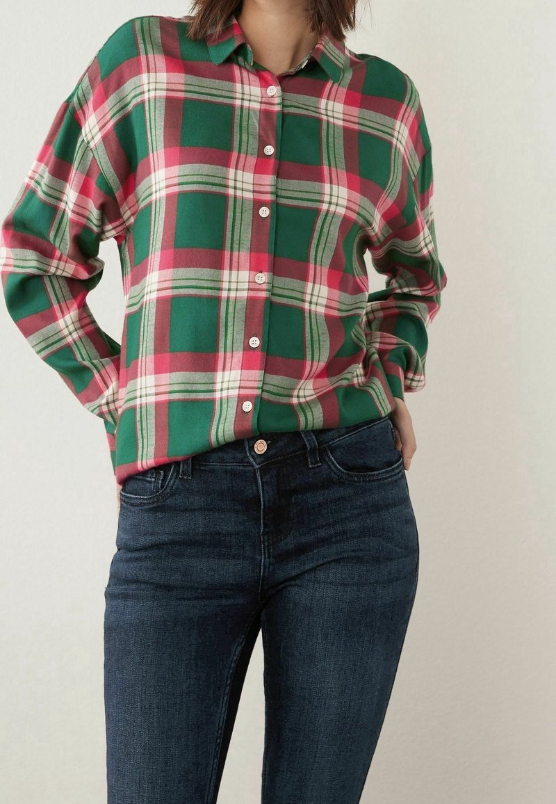 Chemise à carreaux verts et rouges à boutons avec col ; comporte des boutons blancs et des manches retroussées. Associée à un jean slim bleu marine.