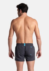 Costume da bagno grigio con elastico in vita, dettagli blu a contrasto e tasca posteriore. Il materiale sembra leggero e liscio.