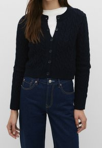 Mörkblå kabelstickad cardigan med ribbade ärmslut och nederkant; har knappstängning, rund halslinje och strukturerat mönster. Matchas med mörka denimjeans.