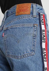 Jeans in denim azzurro chiaro con un'etichetta in pelle con marchio, una linguetta rossa sulla tasca e una banda laterale rossa, bianca e blu navy.