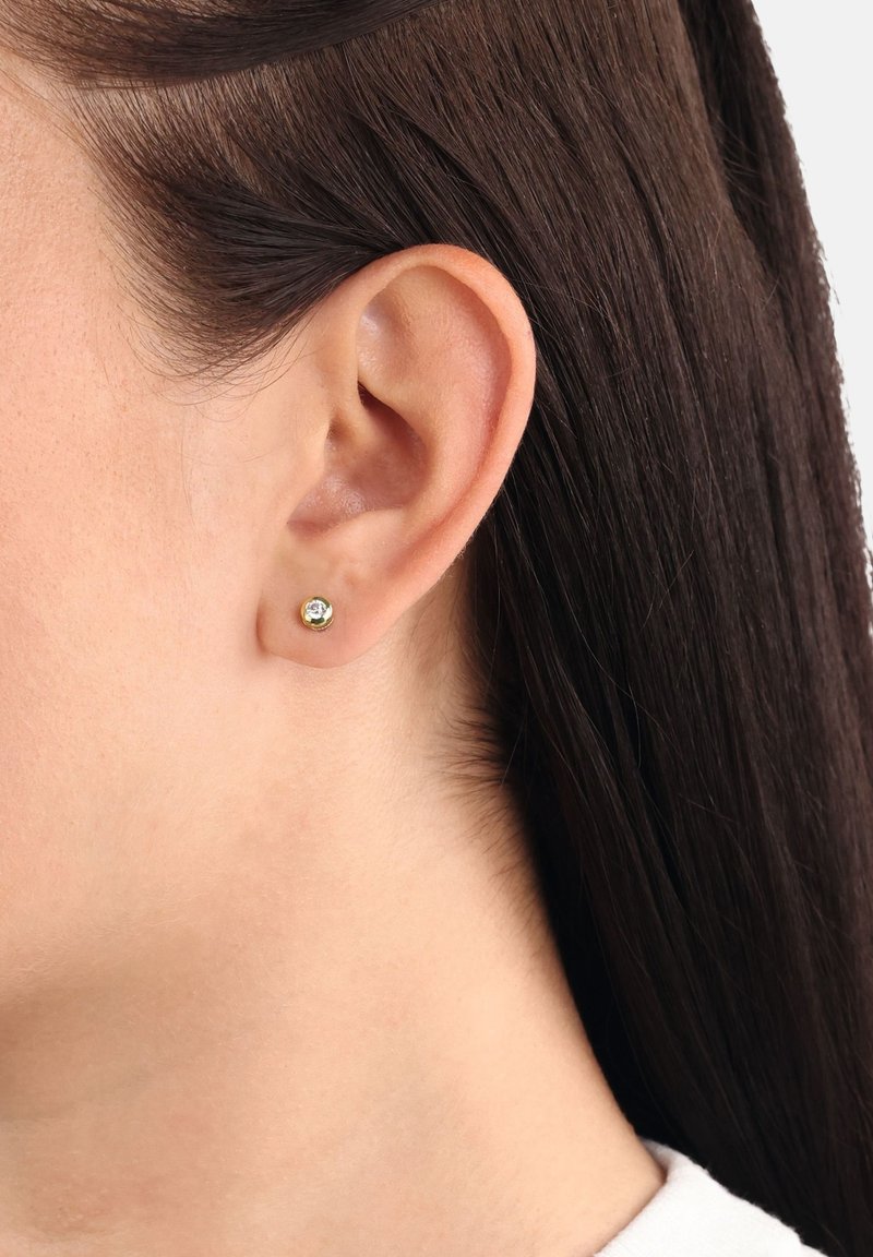 Kvinna med mörkbrunt hår som är bakåttyst, bär en liten rund guldfärgad stiftörhänge med en klar ädelsten i mitten.