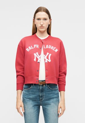 Mladá žena s rovným vlasmi nosí červenú zapínanú bundu Ralph Lauren, bielu košeľu, modré džínsy a hnedý opasok na jednoduchom pozadí.