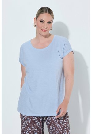 Frau mit hellblauem kurzärmligem Hemd und gemusterten braun-weißen Hosen, stehend vor einem einfachen hellen Hintergrund.