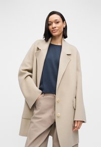 Beige oversized coat with wide sleeves, front pockets, and two buttons, worn over a dark blue shirt and beige trousers. Smooth texture.