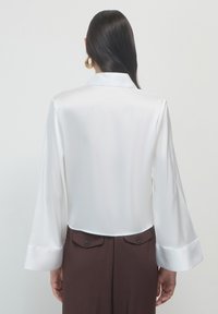 Una donna con i capelli lunghi e scuri indossa una blusa bianca di raso larga con ampi polsini e pantaloni marroni scuri con tasche posteriori con bottoni.