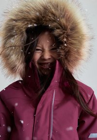 Tyttö hymyilee silmät suljettuina, päällä maroon-värinen talvitakki isolla turkisreunuksella, ympärillä leijuvat lumihiutaleet.