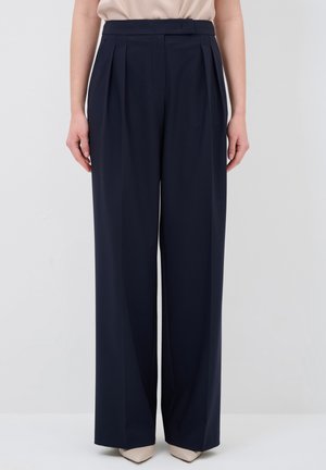 Pantaloni a gamba larga blu navy con pieghe, una texture liscia, vita alta e tasche laterali, abbinati a scarpe ai piedi appuntiti di colore chiaro.